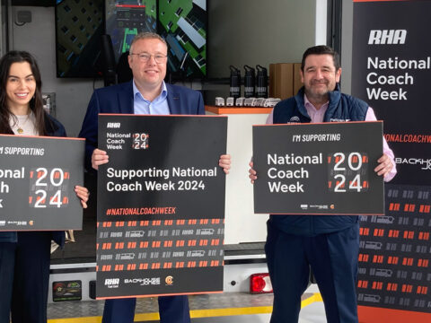 Amelia, Simon and Lee hold up boards supporting National Coach Week 2024 in front of the RHA coach simulator in the Pelican Bus and Coach showroom